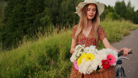 Middle-plan-a-young-sexy-blonde-on-a-bike-in-a-dress-and-flowers-in-a-basket-on-the-handlebar-goes-across-the-field-in-the-summer-walking-on-the-grass-in-slow-motion.